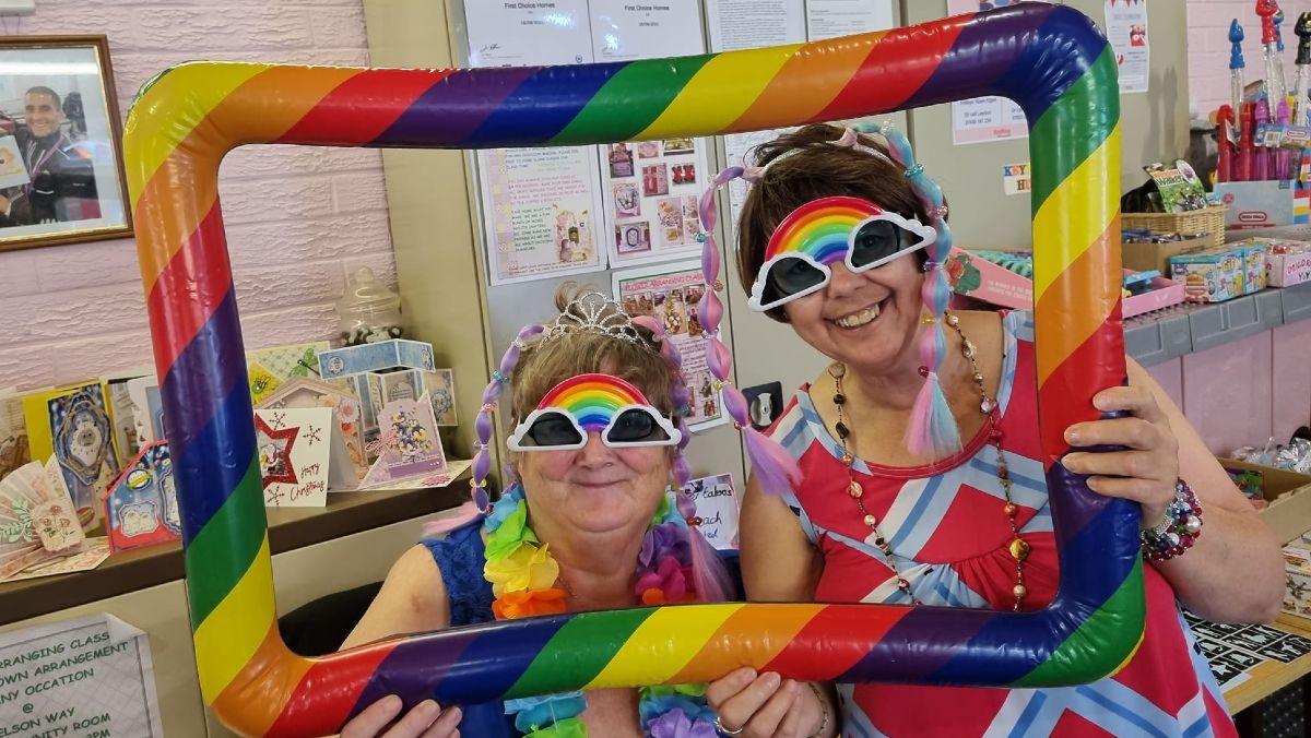 A photo of two women holding up a large rainbow frame and looking through it. They are wearing novelty sunglasses with clouds round the eyes and a rainbow across the top, and colourful clothing.