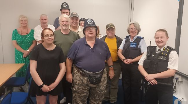 A group of Members standing with two police officers in uniform. Some of the Members are also wearing police hats. It's a jolly picture from a meeting.