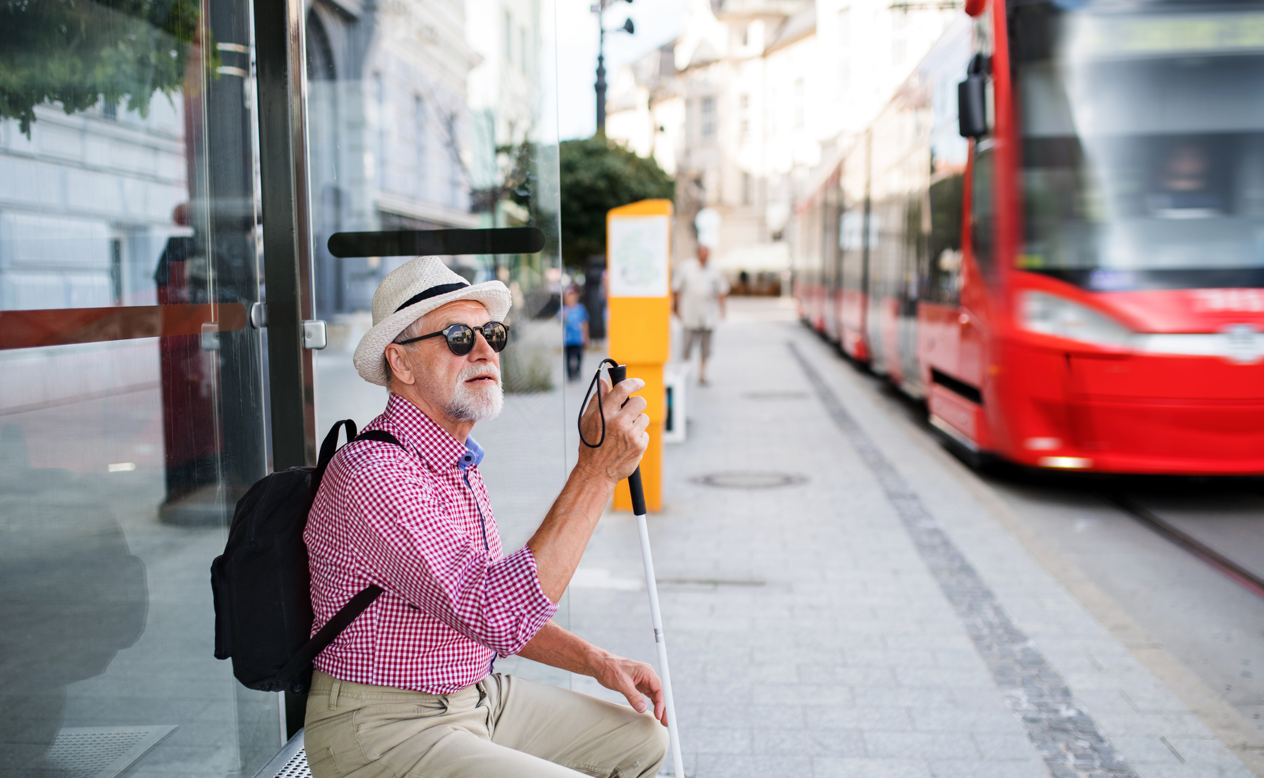 A visually impaired man holding his stick, sitting at a tram stop as a tramp approaches